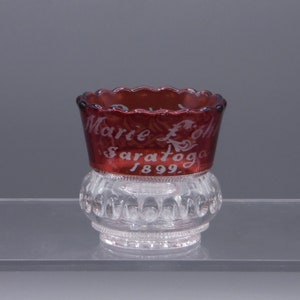 May include: A small, clear glass cup with a red glass top. The top has a scalloped edge and is etched with the words "Marie L.  Saratoga 1899."