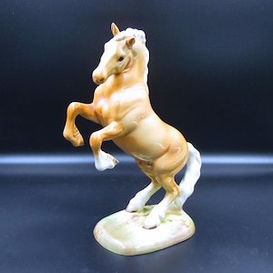 May include: A ceramic figurine of a rearing horse. The horse is a light tan color with a white mane and tail. It stands on a small, oval base with a green and brown color scheme. The horse is posed with its front legs raised.