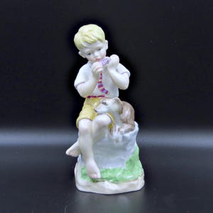 May include: A porcelain figurine of a young child playing a musical instrument, with a dog resting beside him. The child is wearing a white shirt, yellow shorts, and a colorful striped tie. The figurine is set on a rock-like base with green grass.
