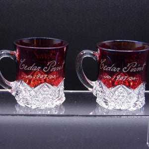 May include: Two red glass souvenir mugs with a clear, cut-glass base. The mugs are engraved with "Cedar Point" and "Est. 1907".