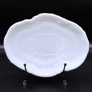 May include: A white, oval-shaped serving dish with a decorative, embossed floral and scroll design. The dish is supported by a black metal stand, and the background is black.