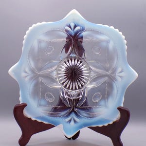 May include: A blue opalescent glass bowl with a scalloped edge and a delicate floral design. The bowl is on a wooden stand.