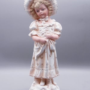 May include: A porcelain figurine of a young girl wearing a white dress and a large white hat. She is holding a bouquet of flowers and has a sweet expression on her face.