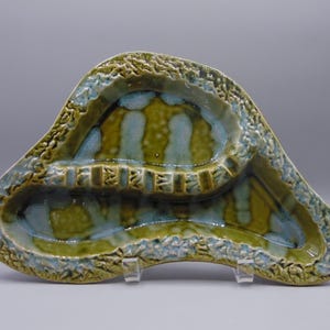 May include: A green and blue ceramic ashtray with a unique, organic shape and three compartments. The ashtray has a textured surface and a glazed finish.