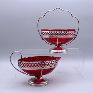 May include: Two-piece set of vintage red glass serving dishes. The pieces have a silver-colored metal filigree around the rim and a silver-colored metal base. One piece has a handle, and the other has a spout.