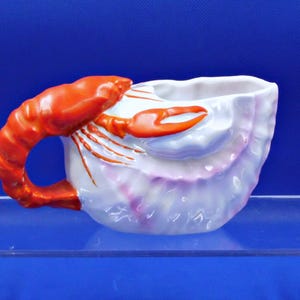 May include: A ceramic mug shaped like a white seashell with a textured, wavy surface and a soft purple tint. The handle is formed by a bright orange lobster, with its body curving to create the grip and its claws resting on the side of the mug.