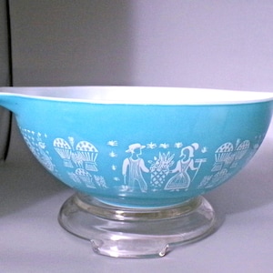 May include: A turquoise Pyrex mixing bowl with a white interior and a clear glass stand. The bowl features a white pattern of figures and baskets. The bowl has a capacity of approximately 4 quarts.