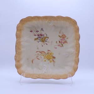 May include: Square, off-white ceramic plate with a scalloped, gold-colored rim. The plate features hand-painted floral designs in shades of purple, pink, and yellow. The plate is displayed on a clear stand.