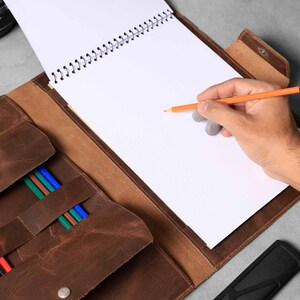 Puede incluir: Un cuaderno de bocetos de cuero marrón con un porta lápices incorporado. El cuaderno está abierto y una mano está dibujando con un lápiz naranja en el papel blanco en blanco.