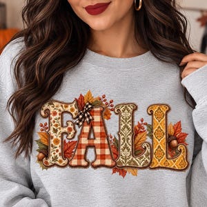 Op de afbeelding: Een lichtgrijs sweatshirt met ronde hals en het woord FALL in grote, decoratieve letters. Elke letter heeft een uniek patroon, waaronder bloemen, rood-wit ruitjespatroon met een klein strikje en ingewikkelde gouden damastontwerpen.