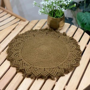 May include: A round, brown macrame placemat with a fringed edge sits on a wooden table. The placemat has a detailed, circular design. A gold vase with white flowers is in the background.