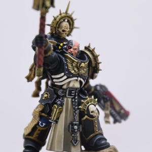 May include: A painted miniature figurine of a space warrior in black and gold armor with a cream-colored tabard. The figure features a bionic eye, a skull-topped staff held aloft, a winged chest emblem, and a skull icon on the knee armor.