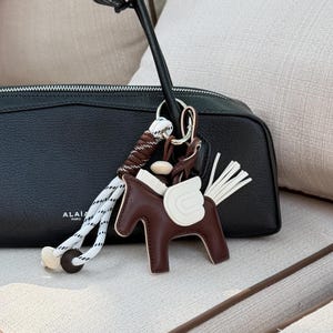 Può includere: Una borsa in pelle nera con la scritta ALAIA PARIS stampata in bianco, dotata di un ciondolo a forma di cavallo marrone e bianco. Il ciondolo presenta un corpo di cavallo in pelle marrone, ali bianche in pelle, una coda a frange e un cordino.