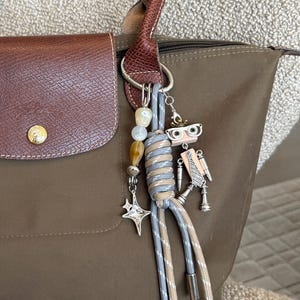 May include: An olive green tote bag with brown leather accents. The bag features a brown flap closure and handles. A decorative charm with beads, a star, and a robot-like figure hangs from the handle.