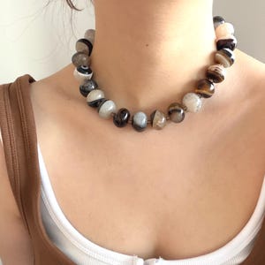 以下が含まれることがあります： 茶色、グレー、白の色合いの、大きくて丸い研磨された石を使ったビーズネックレス。 ネックレスは首の周りに着用されています。 背景は茶色のトップスと白いアンダーシャツです。