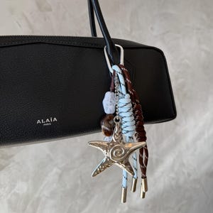 May include: Black leather handbag with a silver starfish charm, brown and blue rope accents, and the brand name "ALAÏA PARIS". The bag has a long, thin strap and a silver clasp.