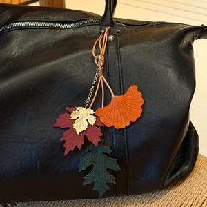 May include: A black leather handbag featuring a decorative charm attached to the handle. The charm consists of three leaf-shaped pendants in dark red, dark green, and orange, along with a metallic gold-coloured maple leaf accent on a chain.