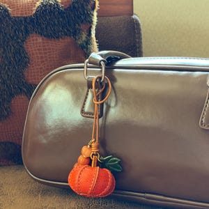 May include: A pumpkin-shaped charm hanging from a brown leather cord, featuring a textured orange pumpkin, two small orange beads, and a green leaf accent. The charm is attached to the handle of a smooth, dark brown handbag with a glossy finish.