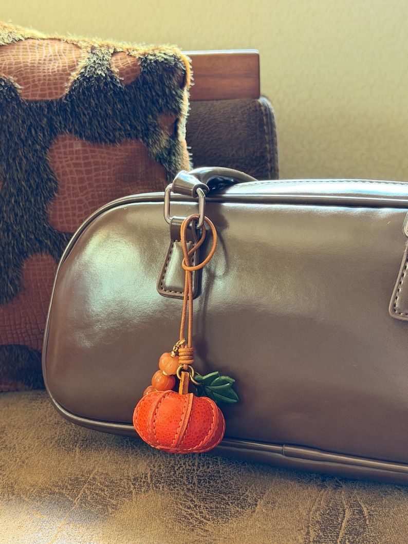 May include: A pumpkin-shaped charm hanging from a brown leather cord, featuring a textured orange pumpkin, two small orange beads, and a green leaf accent. The charm is attached to the handle of a smooth, dark brown handbag with a glossy finish.
