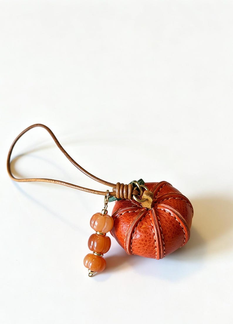 May include: A small, round, orange textured leather pumpkin charm with visible stitching, attached to a thin brown leather cord. A dangling accent features three small, ribbed, translucent orange beads separated by tiny gold-toned metal spacers.