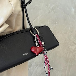 Peut inclure: Sac &agrave; main en cuir noir avec un charm en forme de c&oelig;ur rouge. Le charm est attach&eacute; &agrave; un anneau argent&eacute; et comporte un c&oelig;ur rouge, des perles noires et un cordon ray&eacute; rouge et blanc. Le sac porte la marque "ALA&Iuml;A PARIS".