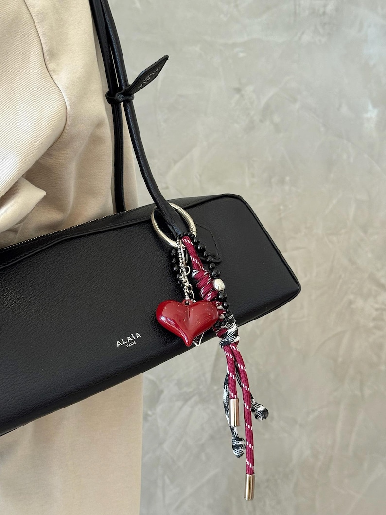 Peut inclure: Sac &agrave; main en cuir noir avec un charm en forme de c&oelig;ur rouge. Le charm est attach&eacute; &agrave; un anneau argent&eacute; et comporte un c&oelig;ur rouge, des perles noires et un cordon ray&eacute; rouge et blanc. Le sac porte la marque "ALA&Iuml;A PARIS".