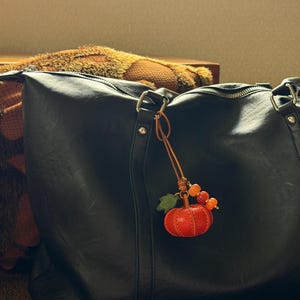 May include: A black leather tote bag featuring a dangling charm accessory. The charm consists of a small, textured orange pumpkin, a green leaf, and three small orange beads attached to the bag's metal hardware by a thin brown leather cord.