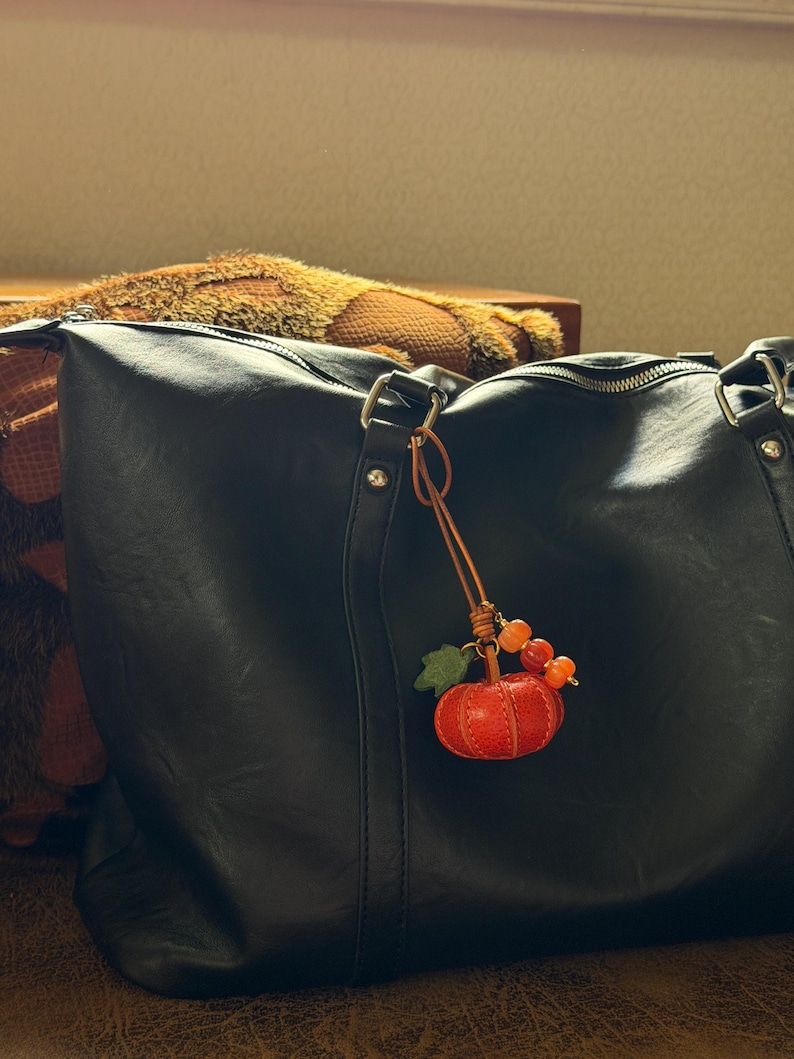 May include: A black leather tote bag featuring a dangling charm accessory. The charm consists of a small, textured orange pumpkin, a green leaf, and three small orange beads attached to the bag's metal hardware by a thin brown leather cord.