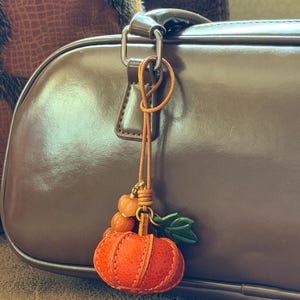 May include: A leather pumpkin bag charm featuring a textured orange pumpkin with green leaves, attached to a thin tan leather cord with small orange beads. The charm hangs from the silver-toned metal hardware of a smooth brown leather handbag.