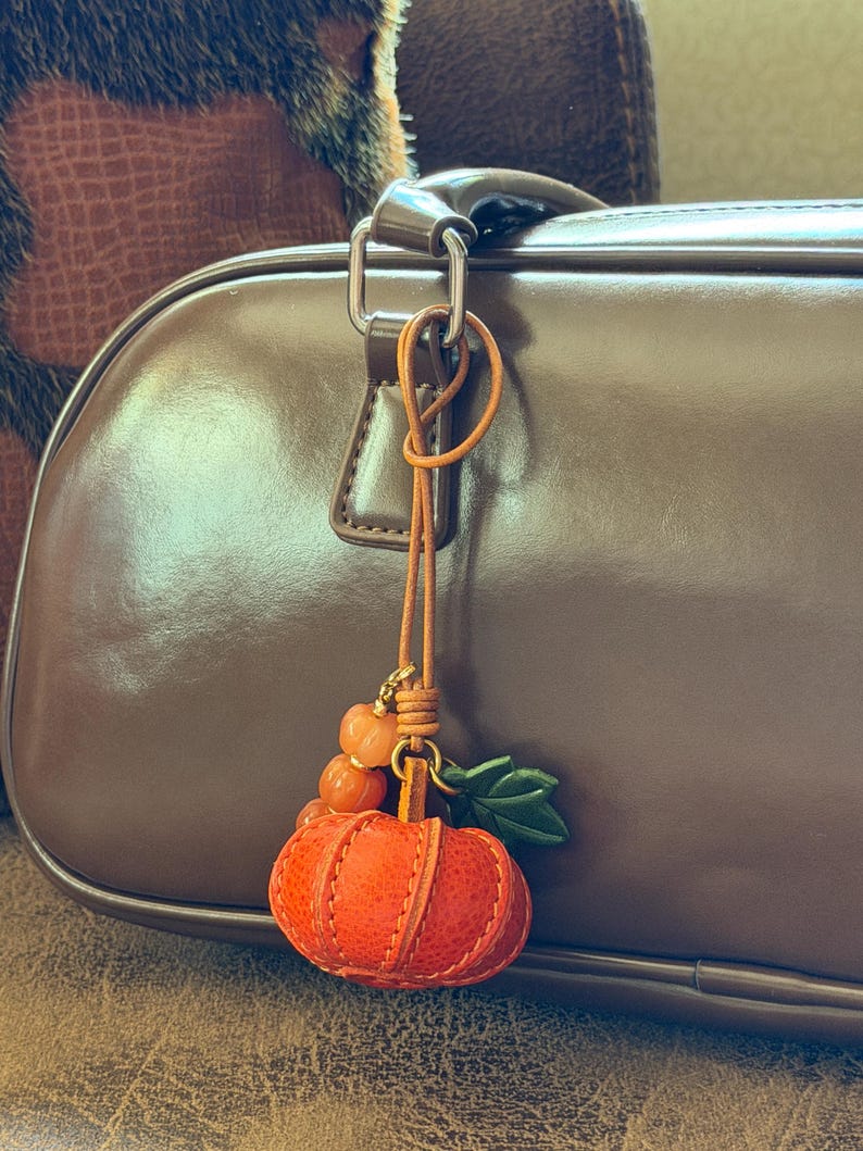 May include: A leather pumpkin bag charm featuring a textured orange pumpkin with green leaves, attached to a thin tan leather cord with small orange beads. The charm hangs from the silver-toned metal hardware of a smooth brown leather handbag.