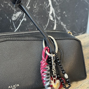Peut inclure: Sac &agrave; main en cuir noir avec une fermeture &eacute;clair argent&eacute;e et un charm d&eacute;coratif. Le charm comprend un c&oelig;ur argent&eacute;, des perles noires et un cordon nou&eacute; rouge, noir et blanc. La marque "ALA&Iuml;A PARIS" est visible.