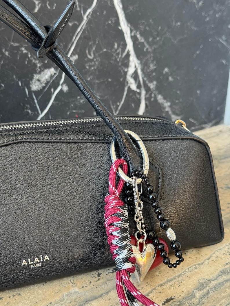 Peut inclure: Sac &agrave; main en cuir noir avec une fermeture &eacute;clair argent&eacute;e et un charm d&eacute;coratif. Le charm comprend un c&oelig;ur argent&eacute;, des perles noires et un cordon nou&eacute; rouge, noir et blanc. La marque "ALA&Iuml;A PARIS" est visible.
