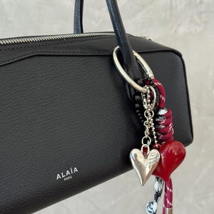 Peut inclure: Sac &agrave; main en cuir noir avec un anneau argent&eacute; et des breloques en forme de c&oelig;ur. Les breloques comprennent un c&oelig;ur argent&eacute; et un c&oelig;ur rouge, attach&eacute;s &agrave; un cordon noir, blanc et rouge.