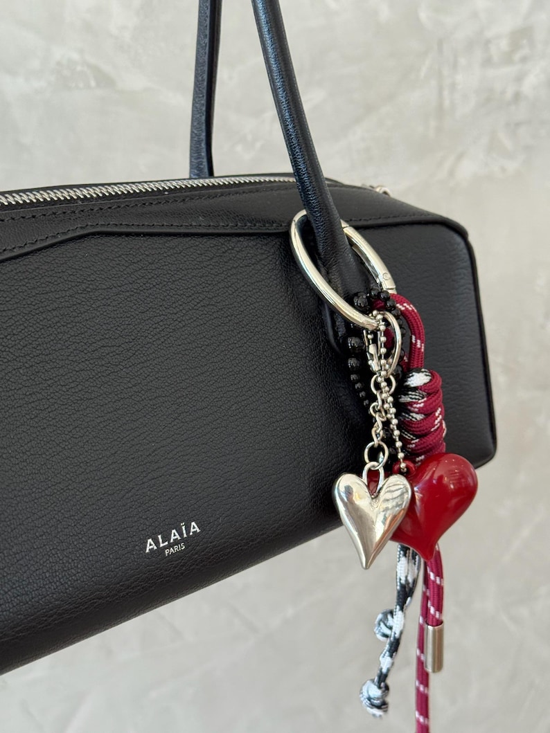 Peut inclure: Sac &agrave; main en cuir noir avec un anneau argent&eacute; et des breloques en forme de c&oelig;ur. Les breloques comprennent un c&oelig;ur argent&eacute; et un c&oelig;ur rouge, attach&eacute;s &agrave; un cordon noir, blanc et rouge.