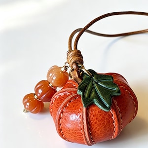 May include: A pumpkin-shaped pendant crafted from textured orange leather with visible stitching, featuring a dark green leather leaf accent. The pendant hangs from a brown leather cord and is decorated with three small, translucent orange beads.