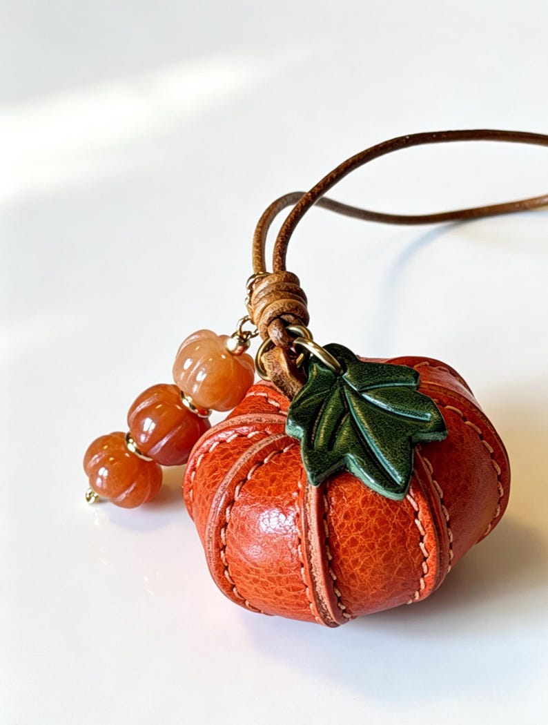 May include: A pumpkin-shaped pendant crafted from textured orange leather with visible stitching, featuring a dark green leather leaf accent. The pendant hangs from a brown leather cord and is decorated with three small, translucent orange beads.