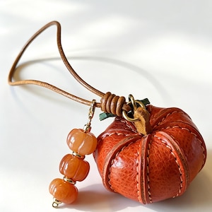 May include: A textured orange leather pumpkin charm attached to a thin brown leather cord. The pumpkin features visible stitching along its segments and a small green leather stem. A gold-toned metal clasp holds three small, ribbed orange beads.