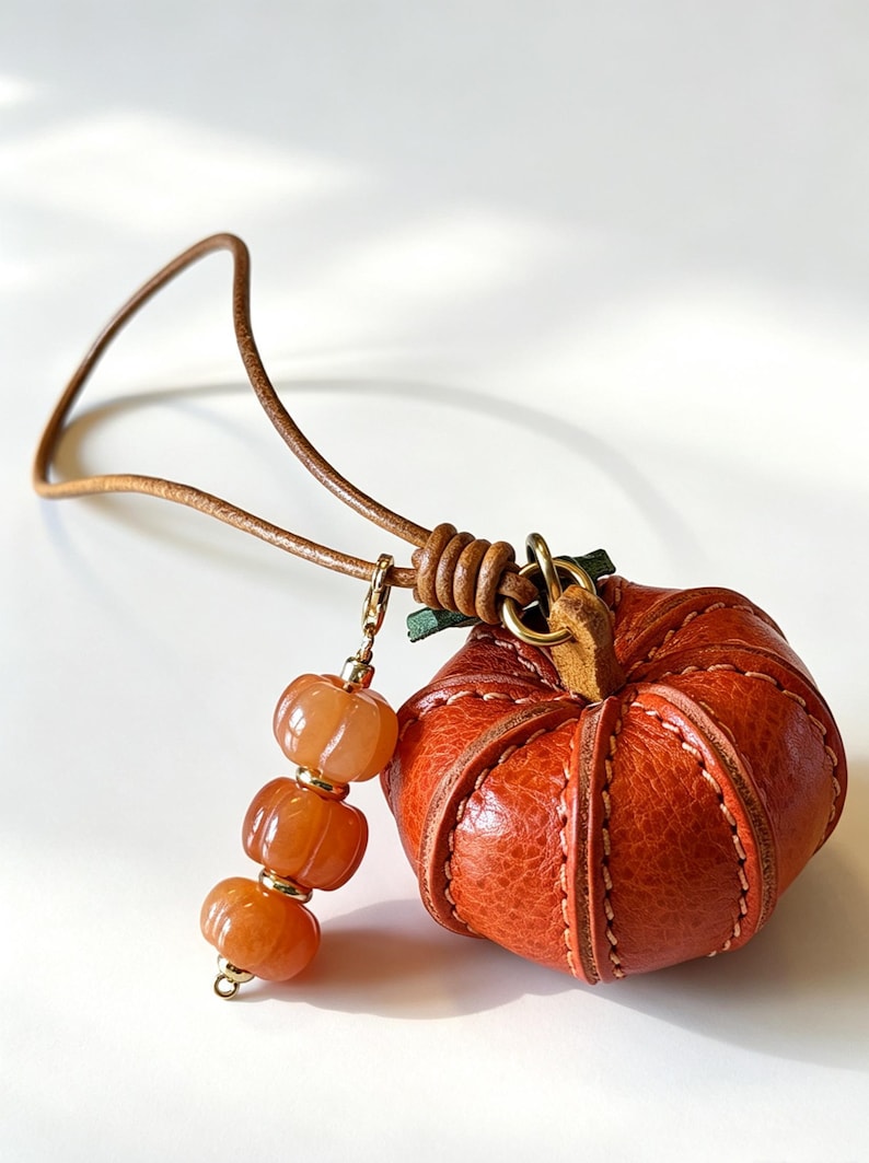 May include: A textured orange leather pumpkin charm attached to a thin brown leather cord. The pumpkin features visible stitching along its segments and a small green leather stem. A gold-toned metal clasp holds three small, ribbed orange beads.