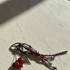 Peut inclure: Porte-cl&eacute;s avec un charm en forme de c&oelig;ur rouge, un charm en forme de c&oelig;ur argent&eacute; et un cordon tress&eacute; rouge, noir et blanc. Le porte-cl&eacute;s a un anneau argent&eacute; et des perles noires.