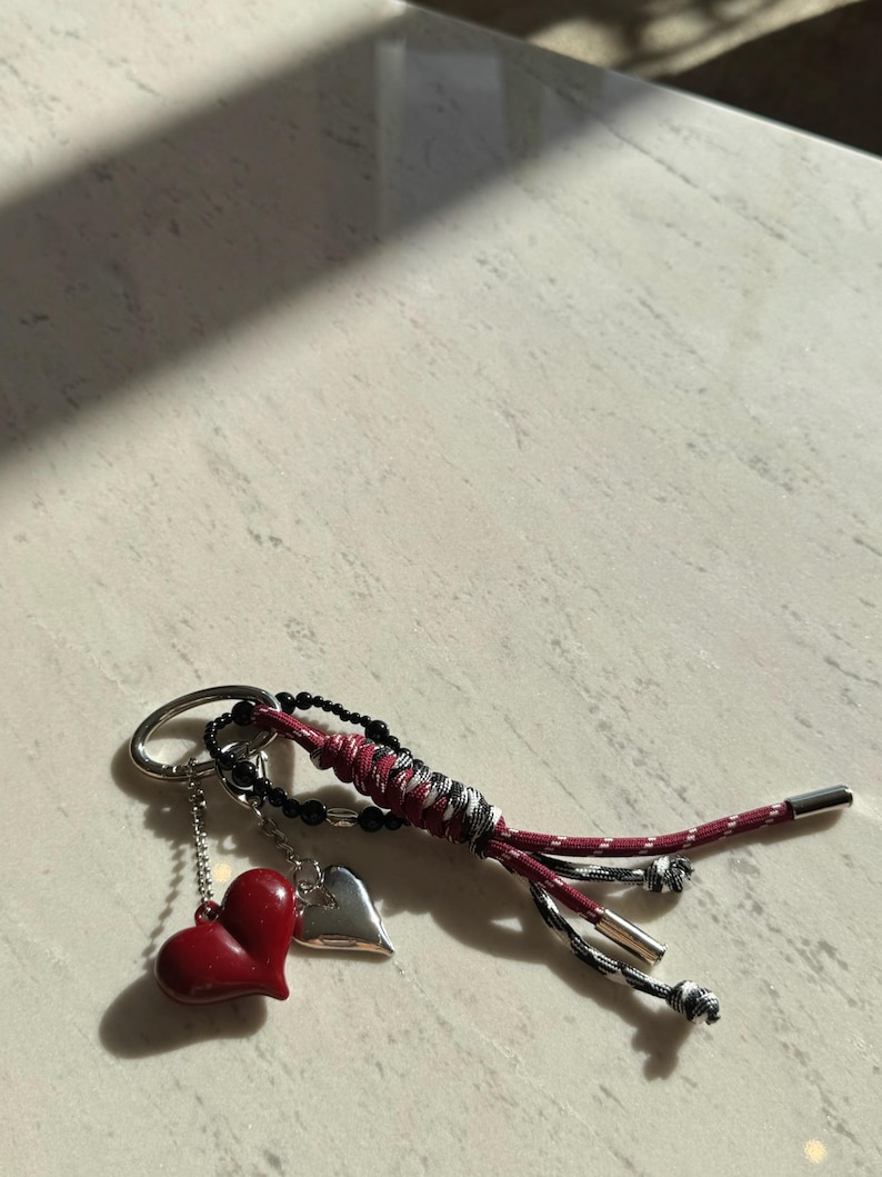 Peut inclure: Porte-cl&eacute;s avec un charm en forme de c&oelig;ur rouge, un charm en forme de c&oelig;ur argent&eacute; et un cordon tress&eacute; rouge, noir et blanc. Le porte-cl&eacute;s a un anneau argent&eacute; et des perles noires.