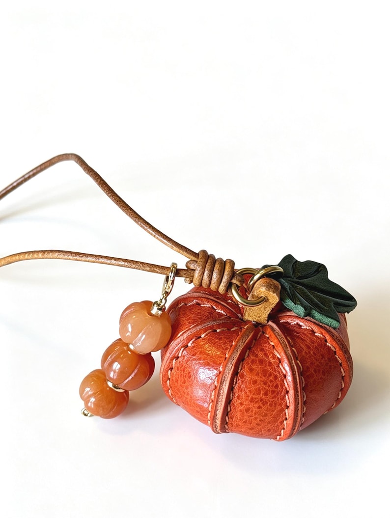 May include: A leather pumpkin pendant on a brown cord, featuring a textured orange leather pumpkin with visible stitching, a dark green leather leaf accent, and a dangling charm consisting of three small, ribbed, translucent orange beads.