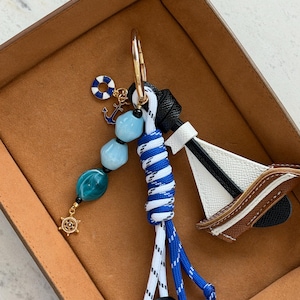 May include: A nautical-themed keychain featuring a small sailboat, blue and white rope, and decorative beads. The keychain includes a gold-colored ring, a life preserver charm, and an anchor charm. The sailboat has white, black, and brown accents.