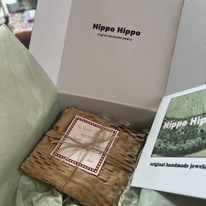 Peut inclure: Une bo&icirc;te blanche ouverte avec le texte "Hippo Hippo original handmade jewelry" contient un paquet emball&eacute;. Le paquet est envelopp&eacute; dans du papier brun et ficel&eacute;. Une carte dit "To Kiasmi, Hope my jewelry brings you happiness, Ash."