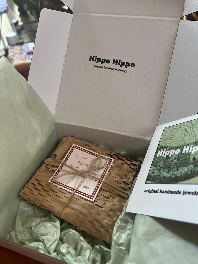 Peut inclure: Une bo&icirc;te blanche ouverte avec le texte "Hippo Hippo original handmade jewelry" contient un paquet emball&eacute;. Le paquet est envelopp&eacute; dans du papier brun et ficel&eacute;. Une carte dit "To Kiasmi, Hope my jewelry brings you happiness, Ash."