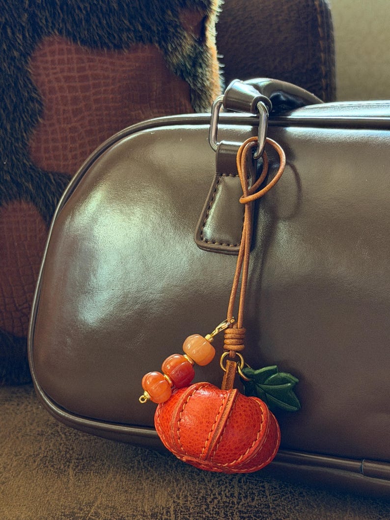 May include: A leather pumpkin charm hanging from a brown handbag. The charm features a textured orange leather pumpkin, a small green leather leaf, and a strand of three smooth, translucent orange beads attached by a thin leather cord.