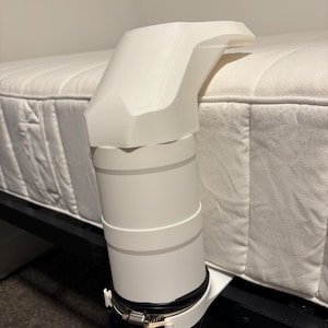 May include: A white 3D-printed plastic vent attachment secured to a mattress edge with a metal hose clamp. The component features a rigid, angled top section connected to a cylindrical base, which is attached to a black flexible corrugated hose.