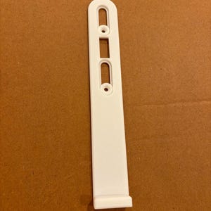 Puede incluir: Un soporte de montaje de pl&aacute;stico blanco con un cuerpo rectangular largo y estrecho con un extremo superior redondeado. El componente incluye dos aberturas verticales ranuradas y dos orificios circulares para tornillos alineados.