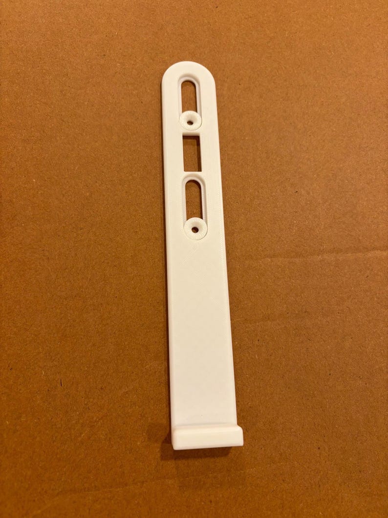 Puede incluir: Un soporte de montaje de pl&aacute;stico blanco con un cuerpo rectangular largo y estrecho con un extremo superior redondeado. El componente incluye dos aberturas verticales ranuradas y dos orificios circulares para tornillos alineados.