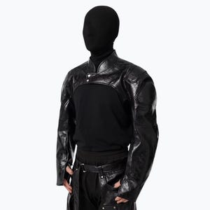 May include: A black faux leather bolero jacket featuring a high collar, long sleeves with structured shoulder panels, and a cropped front design. The garment has a glossy, textured finish and is worn over a plain black long-sleeve shirt.