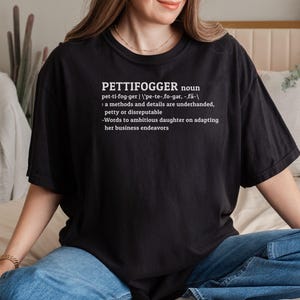 May include: A black t-shirt with white text that defines the word "pettifogger" as a noun. The definition reads: "pet-ti-fog-ger | 'pe-te-,fo-gar, -,fä- : a methods and details are underhanded, petty or disreputable -Words to ambitious daughter on adapting her business endeavors"